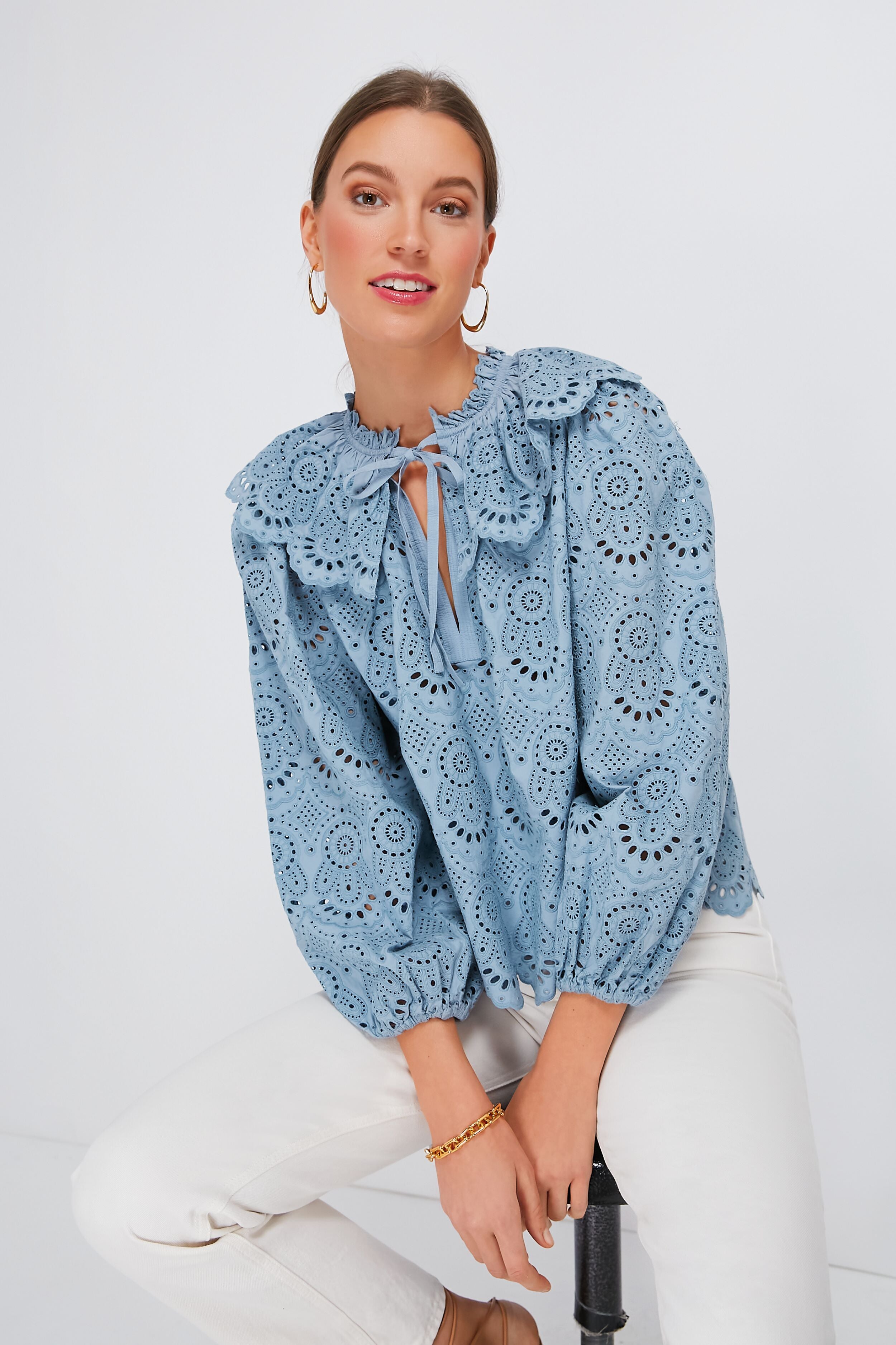 Woman in a light blue Ulla Johnson blouse with eyelet details and a ruffled collar, paired with white pants, seated on a stool.