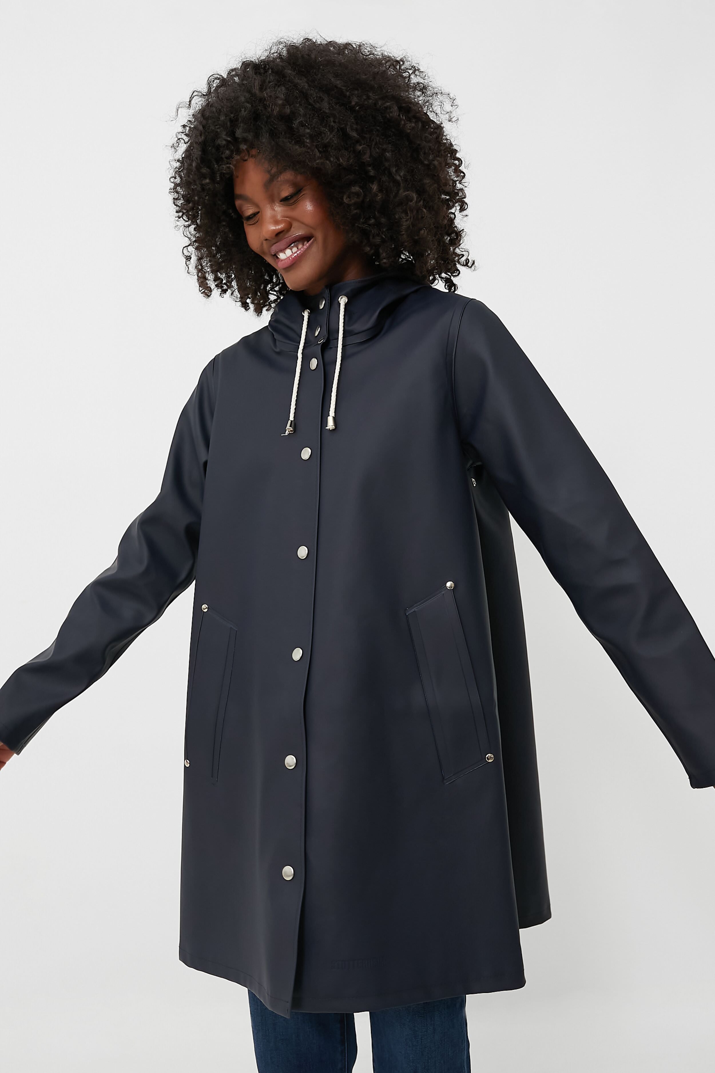 A person wears a navy Stutterheim Mosebacke jacket featuring a hood and snap buttons, smiling with arms slightly outstretched.