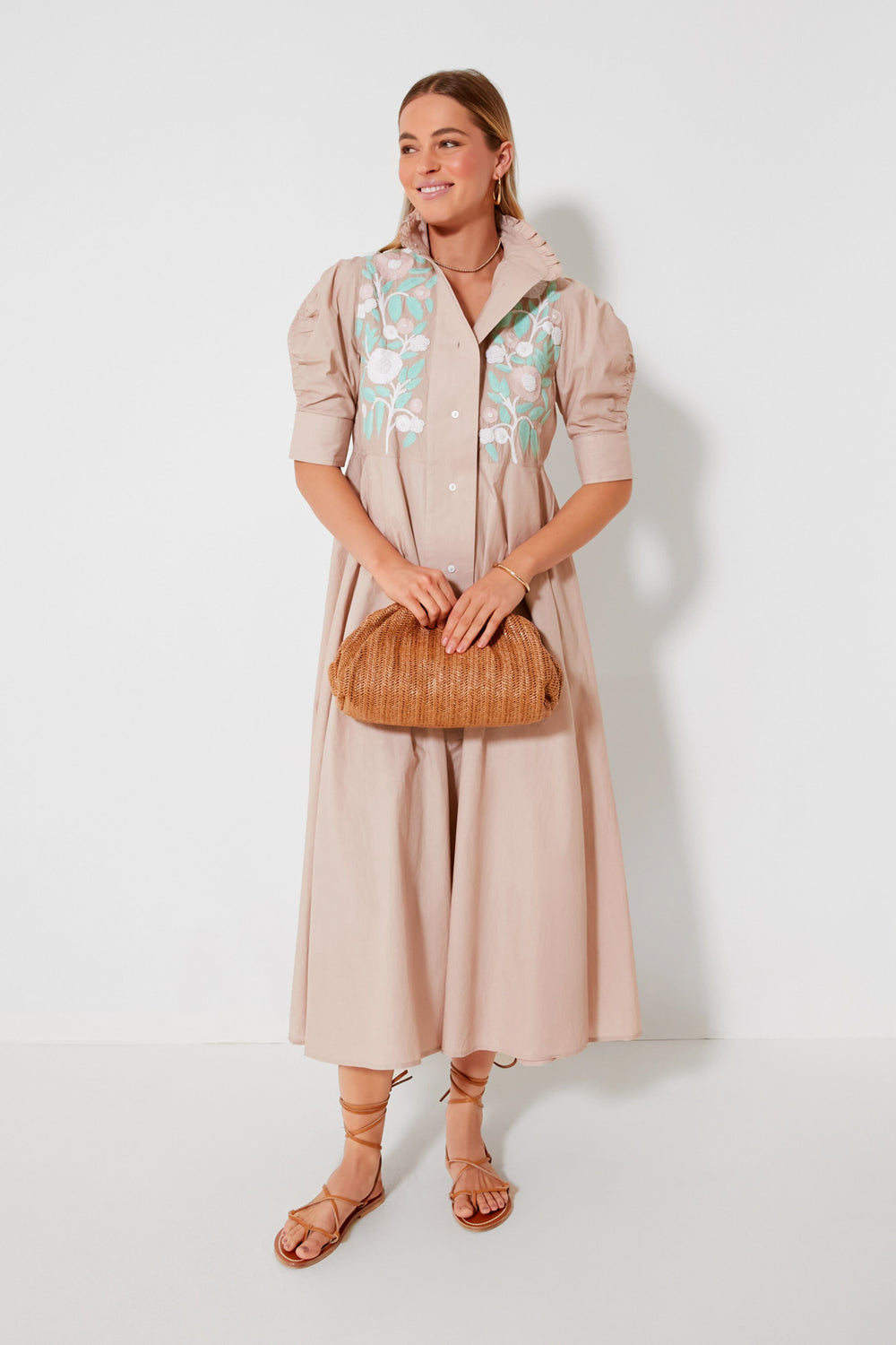 A woman wears a light beige embroidered maxi dress with puff sleeves by Thierry Colson, styled with a woven clutch and brown strappy sandals, evoking Indian blockprint inspiration.
