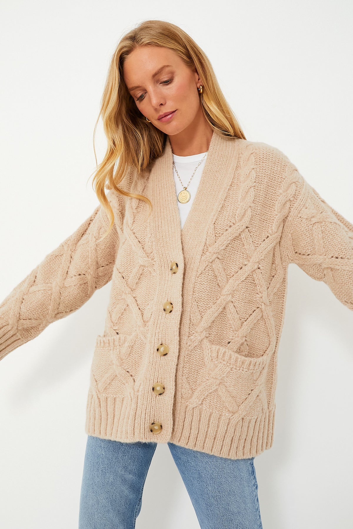 A woman models ELEVEN SIX’s chunky beige Peruvian knit cardigan with front pockets and large buttons, styled with a white top and light blue jeans, channeling classic Andean-inspired style.