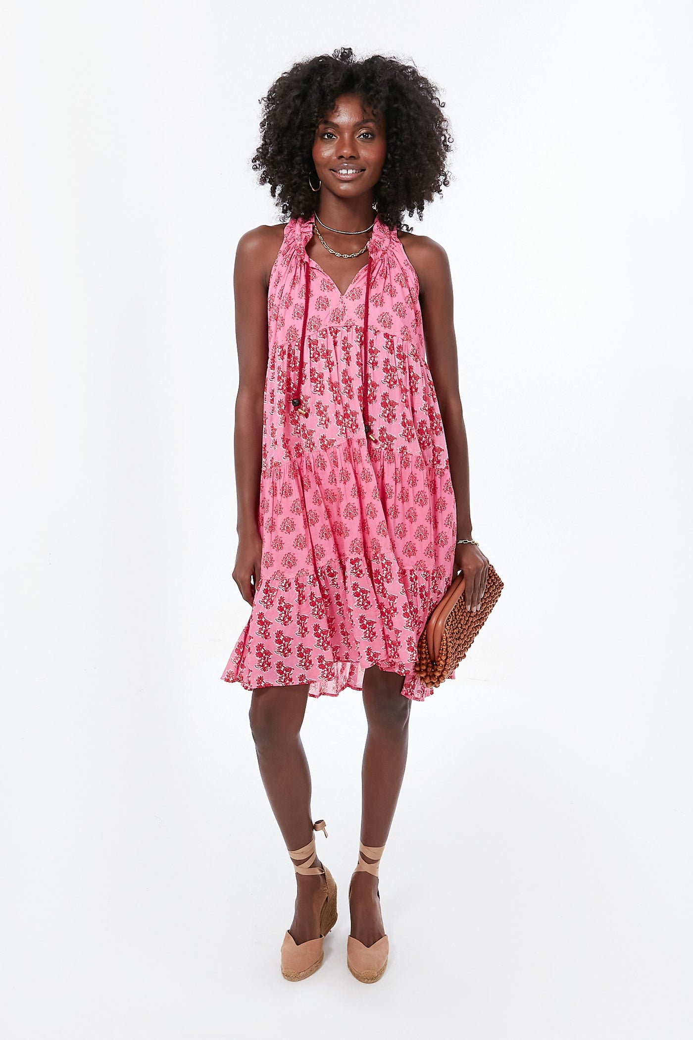 Woman in a Ro’s Garden pink floral halter mini dress, beige wedge sandals, layered necklaces, and holding a brown woven clutch—an ideal luxury travel look.
