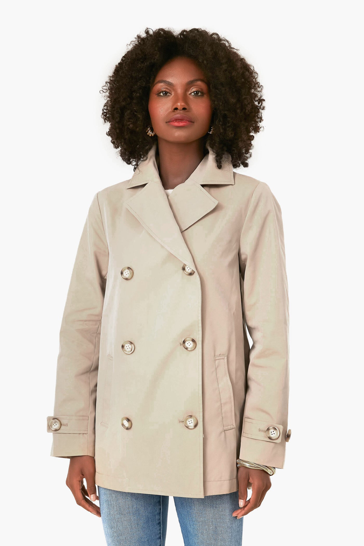 A woman with curly hair wears a light beige water-resistant KULE coat with metallic buttons over blue jeans and a striped tee.