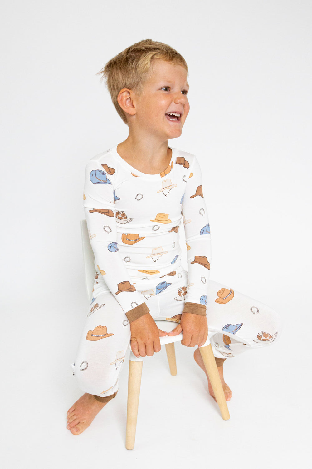 A young boy sits on a small wooden stool, smiling and looking aside in an Angel Dear cozy baby loungewear set made from soft bamboo viscose, featuring a charming cowboy hat print.