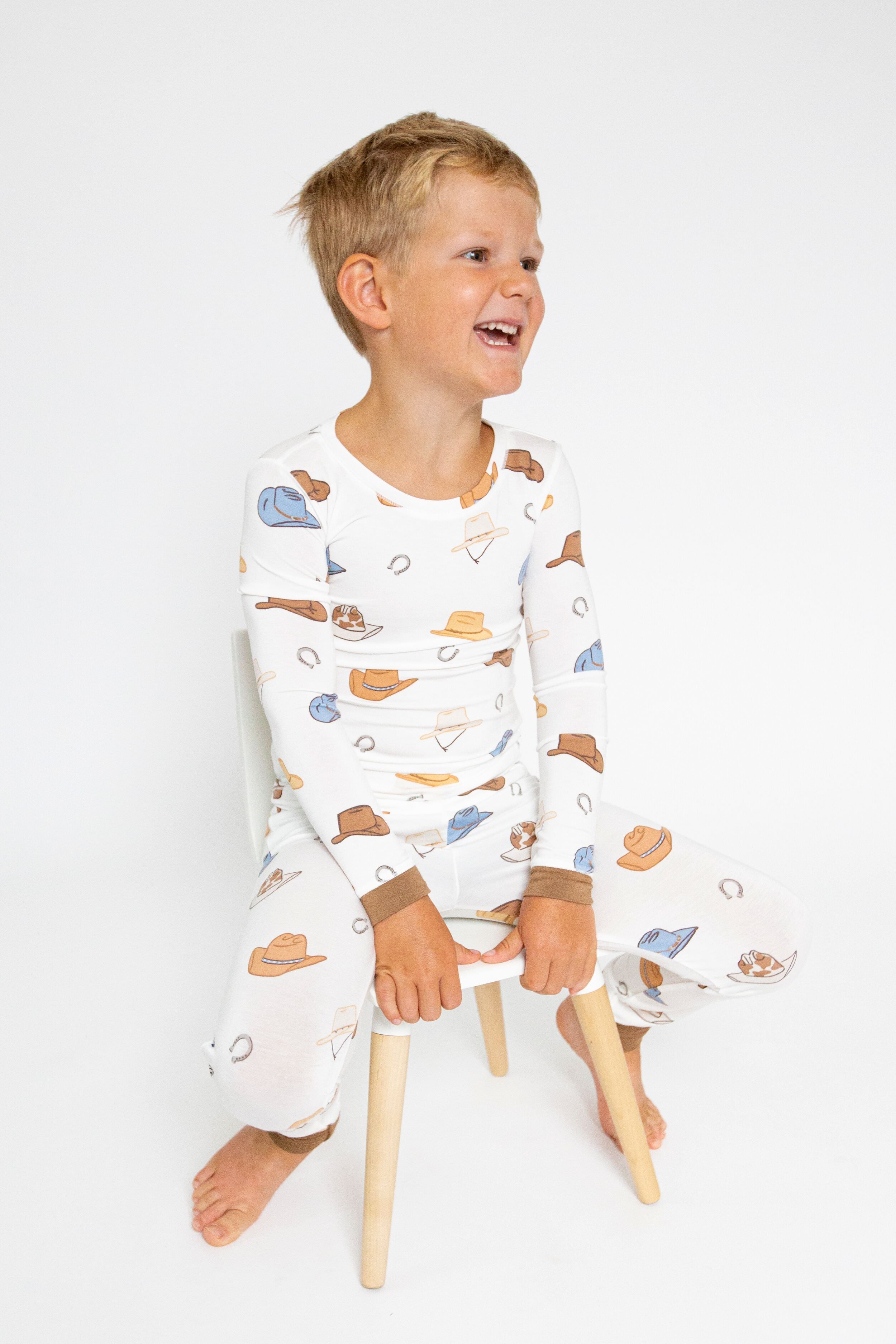A young boy sits on a small wooden stool, smiling and looking aside in an Angel Dear cozy baby loungewear set made from soft bamboo viscose, featuring a charming cowboy hat print.
