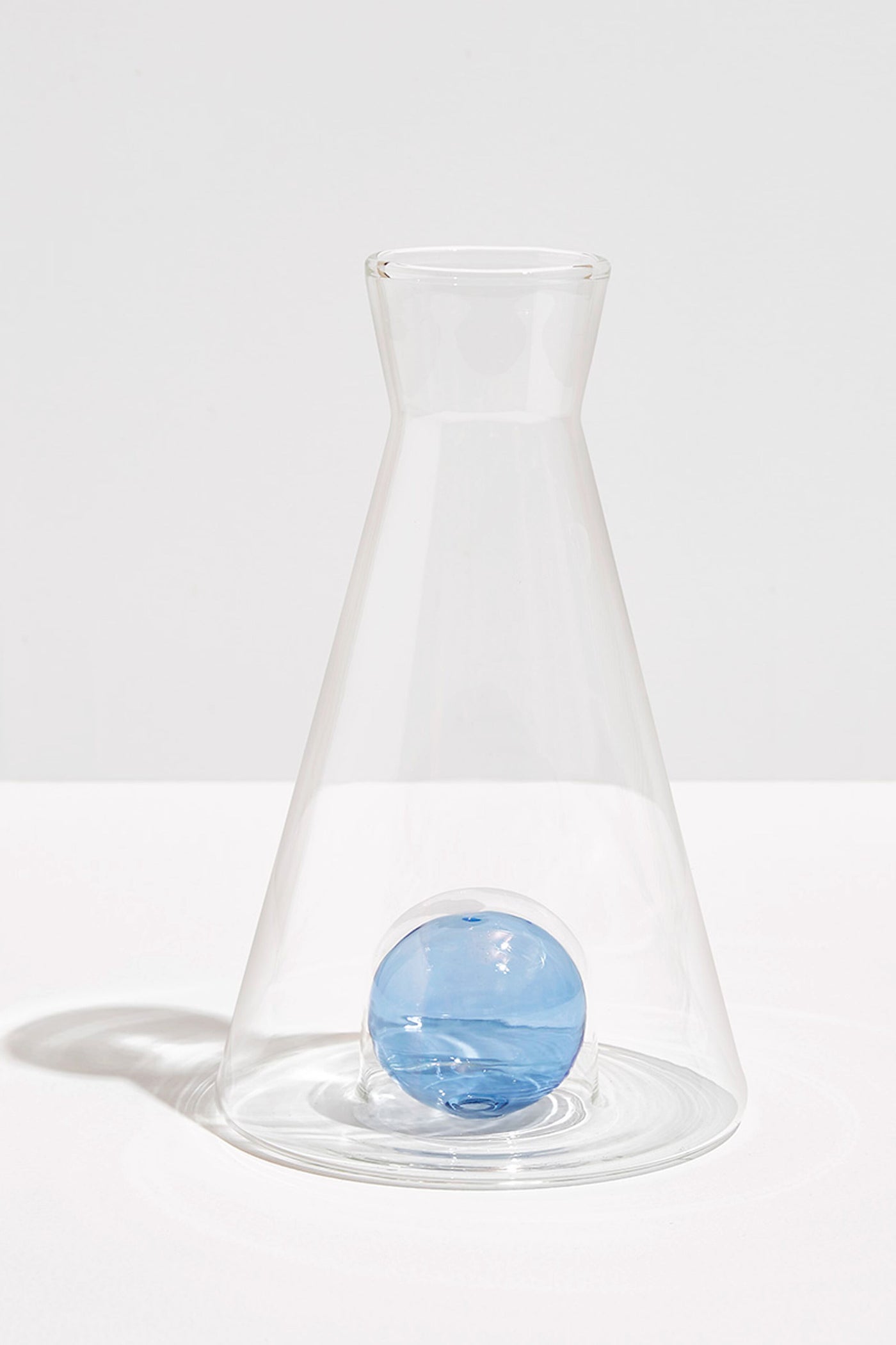 The FAZEEK clear borosilicate glass flask features a wide base, narrow neck, and a single blue glass sphere at the bottom for an elegant sculptural homeware accent.