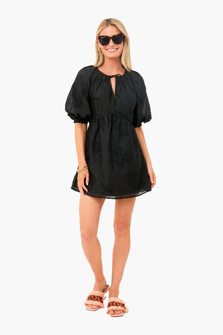 A woman stands smiling in a French-inspired FRAME Noir Eyelet Puff Sleeve Dress with embroidery, sunglasses, and pink ruffle sandals. The dress is made from eco-friendly cotton ramie for effortless chic style.