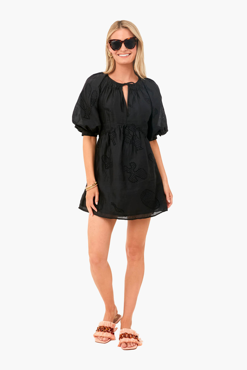 A woman stands smiling in a French-inspired FRAME Noir Eyelet Puff Sleeve Dress with embroidery, sunglasses, and pink ruffle sandals. The dress is made from eco-friendly cotton ramie for effortless chic style.