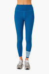 A person is shown from waist to feet wearing Addison Bay high-rise, blue patterned leggings with white stripes near the left ankle and gray sneakers. The leggings feature sweat-wicking fabric for comfortable activewear.