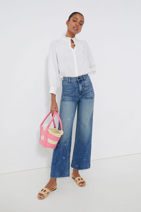 A woman wears a Pomander Place White Emory Smocked Blouse with a smocked bodice and ruffle collar, styled with wide-leg blue jeans, beige sandals, and a pink and beige bucket bag.