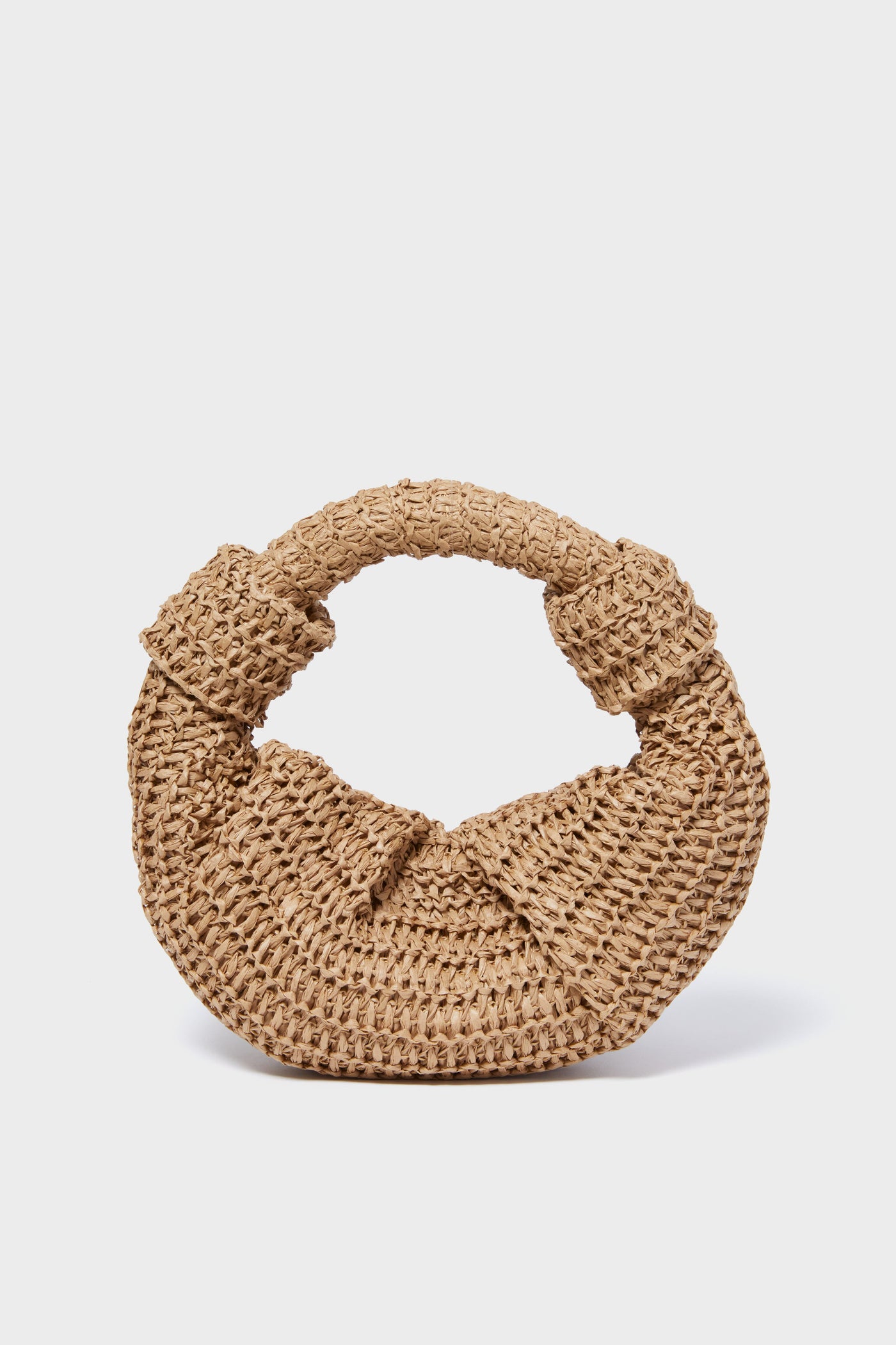 A round raffia bag by Simon Miller, showcasing a thick knot handle and intricate woven details, is displayed in a simple, uncluttered setting.