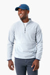 A man wearing a light gray Peter Millar stretch performance quarter-zip sweater, dark pants, and a blue baseball cap stands smiling and looking to the side.