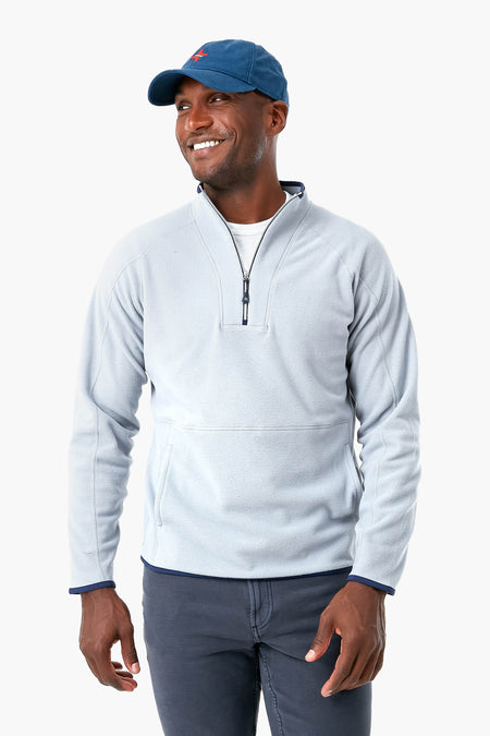 A man wearing a light gray Peter Millar stretch performance quarter-zip sweater, dark pants, and a blue baseball cap stands smiling and looking to the side.