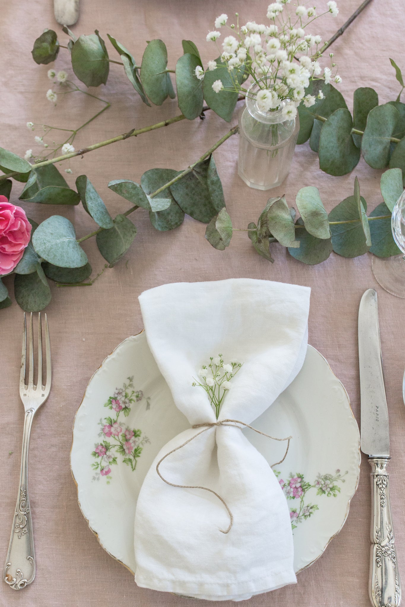 A table is set with a floral plate, fork, and knife. Madame de la Maison Blanc Linen Napkins, tied with twine and small flowers, rest on the plate. Eucalyptus and blooms add charm to these sustainable table linens.