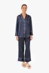 A person smiles while wearing Petite Plume's classic navy blue polka dot pajamas with white piping and cream slippers, embodying effortless luxury sleepwear.