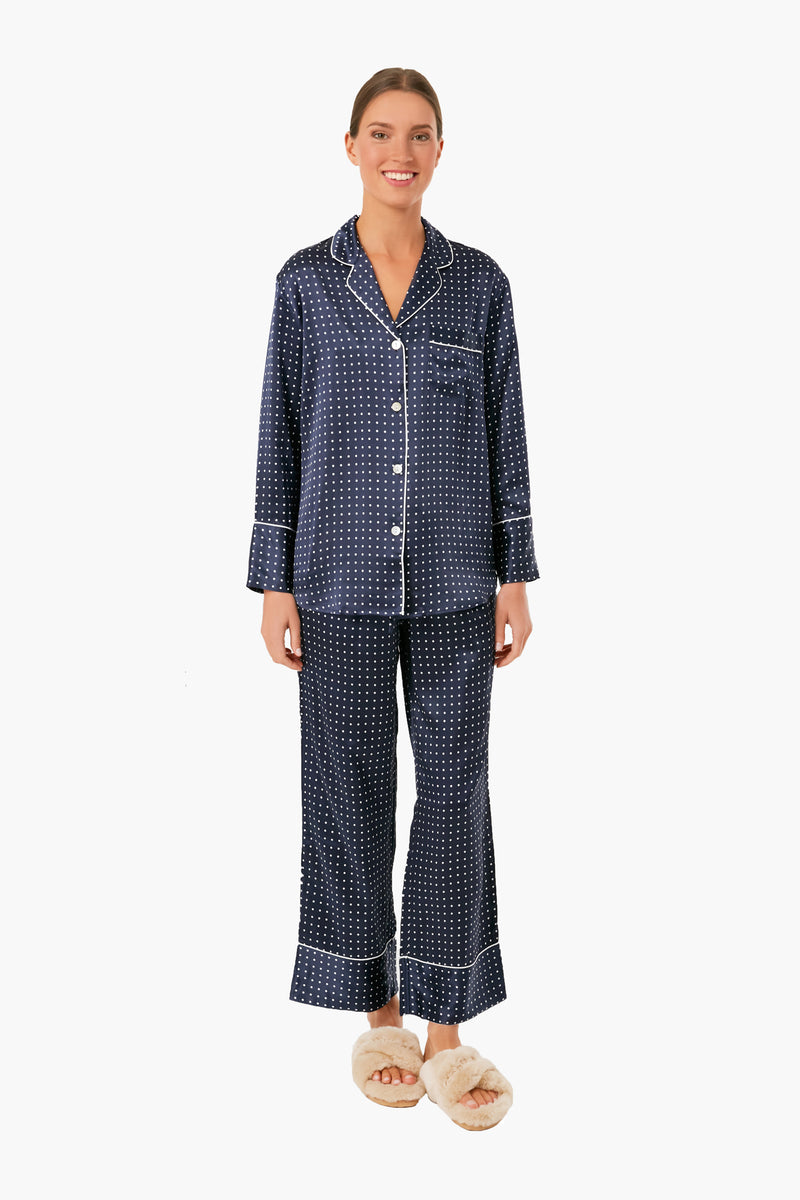 A person smiles while wearing Petite Plume's classic navy blue polka dot pajamas with white piping and cream slippers, embodying effortless luxury sleepwear.