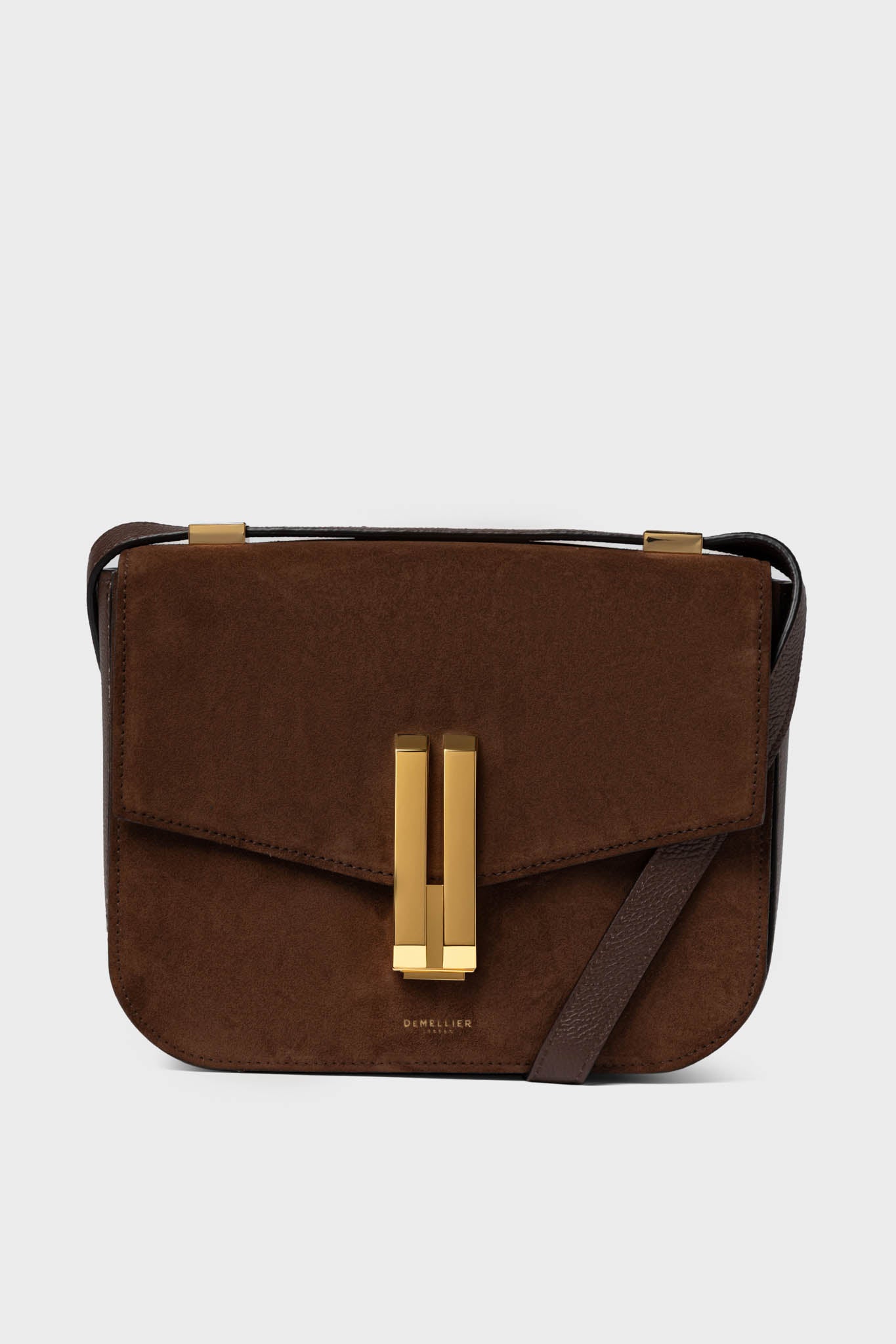 A brown sueded leather crossbody bag by Demellier, featuring a rectangular gold clasp and adjustable strap, capturing the brand’s signature style.