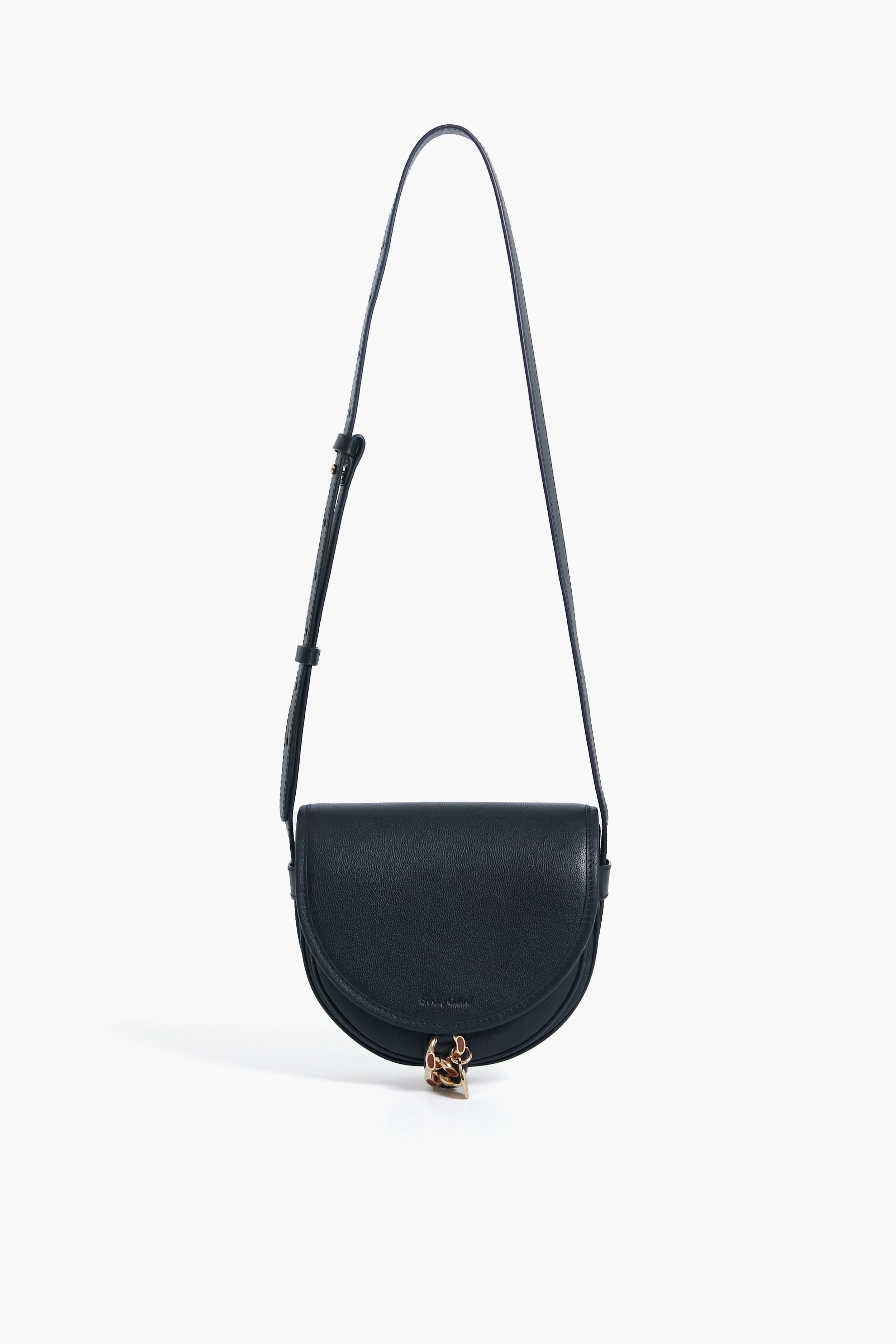 A black See by Chloe crossbody bag made from cowhide, featuring a curved flap, adjustable strap, and gold clasp accent at the bottom center, inspired by the Mara Bag design.