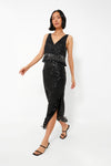 A woman wears Saylor’s sleeveless black sequin dress with fringe, V-neckline, and front slit, paired with black strappy heels—an elegant look for an evening soiree.