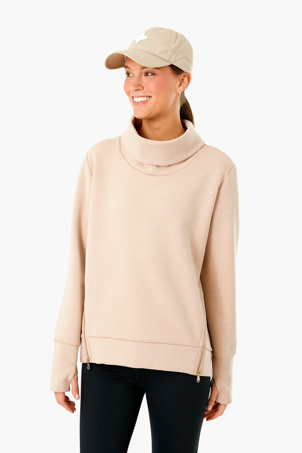 A woman stands and smiles in sporty athleisure wear from Addison Bay, featuring a beige funnel neck sweatshirt, black leggings, and a matching beige baseball cap.