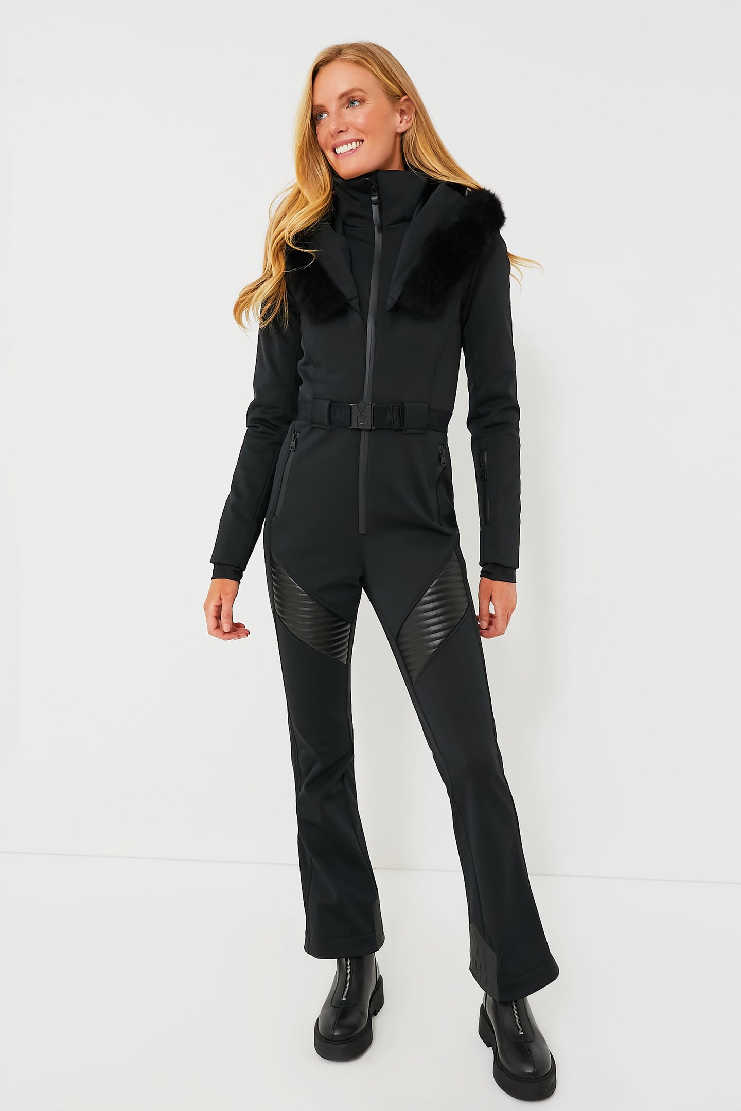 A woman models a MACKAGE black, water-resistant down ski suit with faux fur trim, black boots, and a belted waist.
