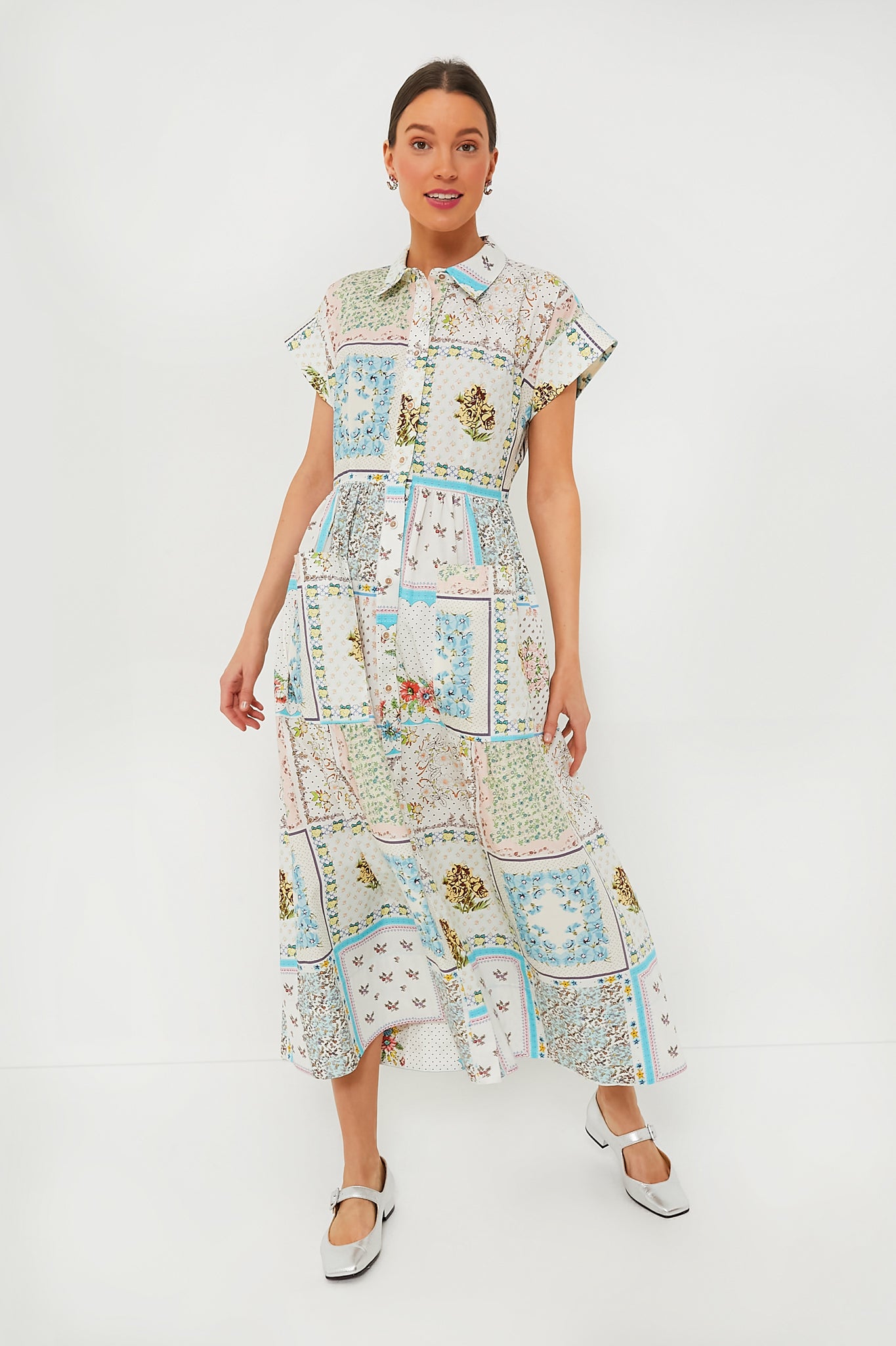 A woman wears the Hunter Bell Patchwork Quilt Sarah Dress, featuring banded cap sleeves, a collared neckline, a tiered skirt, and a floral patchwork print, styled with white shoes.