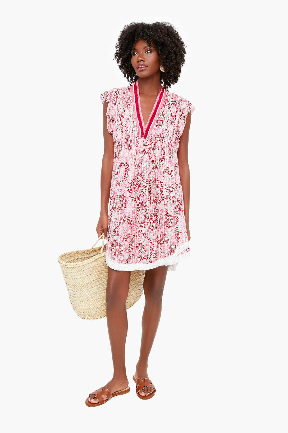 A woman wears a boho-chic, red and white patterned Poupette St Barth dress with cap sleeves, carrying a woven straw tote bag and wearing brown sandals.