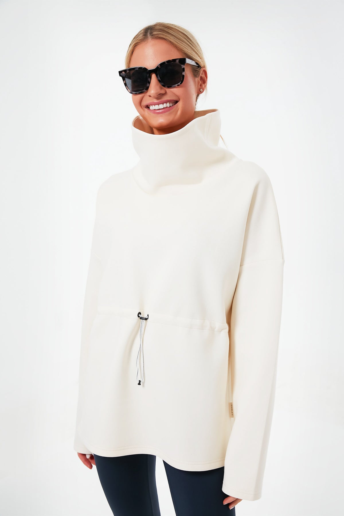 A woman stands and smiles, wearing a Varley Barton Sweat—an oversized funnel collar sweatshirt with a drawstring waist—paired with black leggings and dark sunglasses.