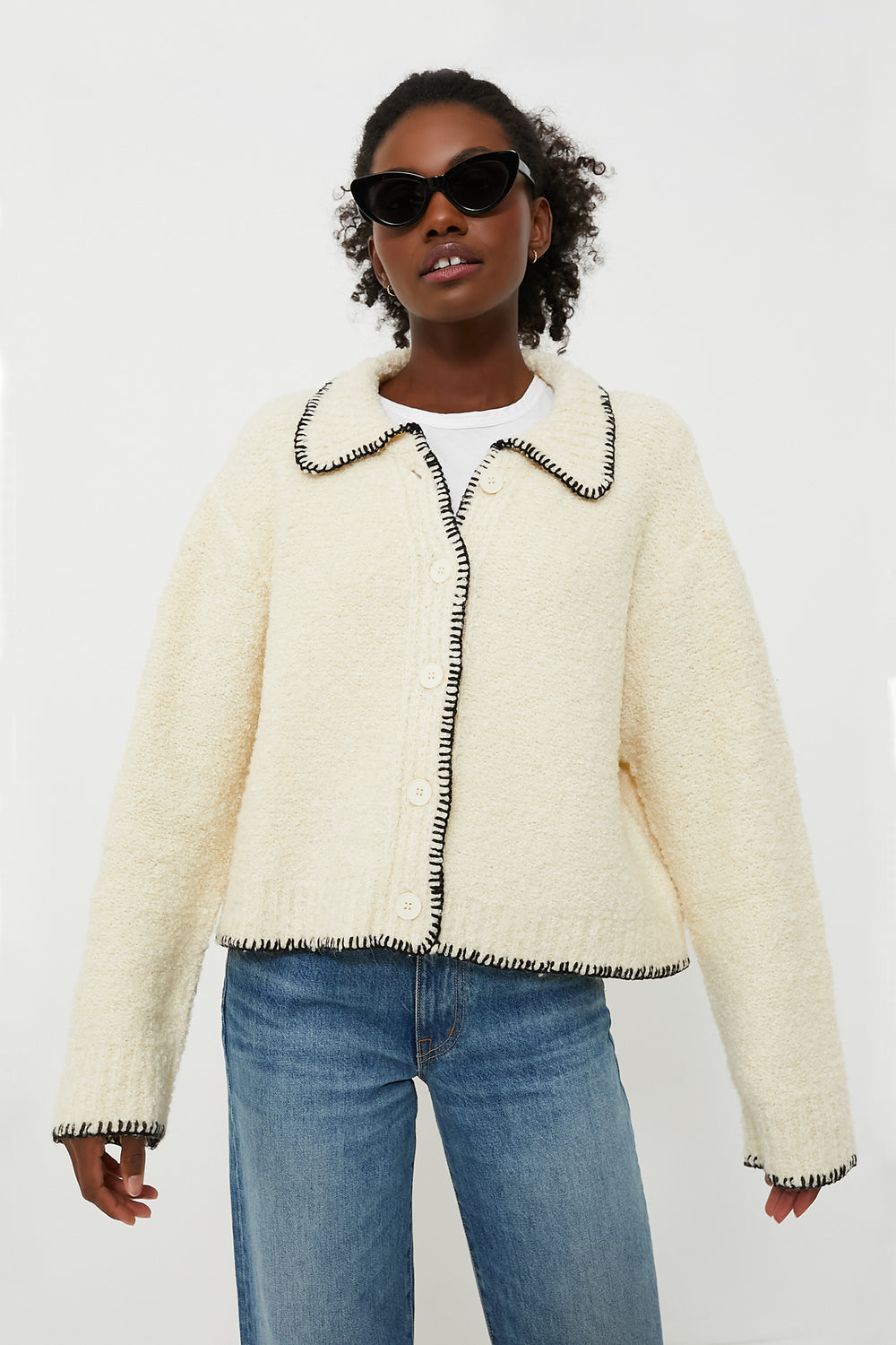 A person wearing black sunglasses, a cream Moon River cardigan with contrast stitching and black trim, and blue jeans stands confidently.