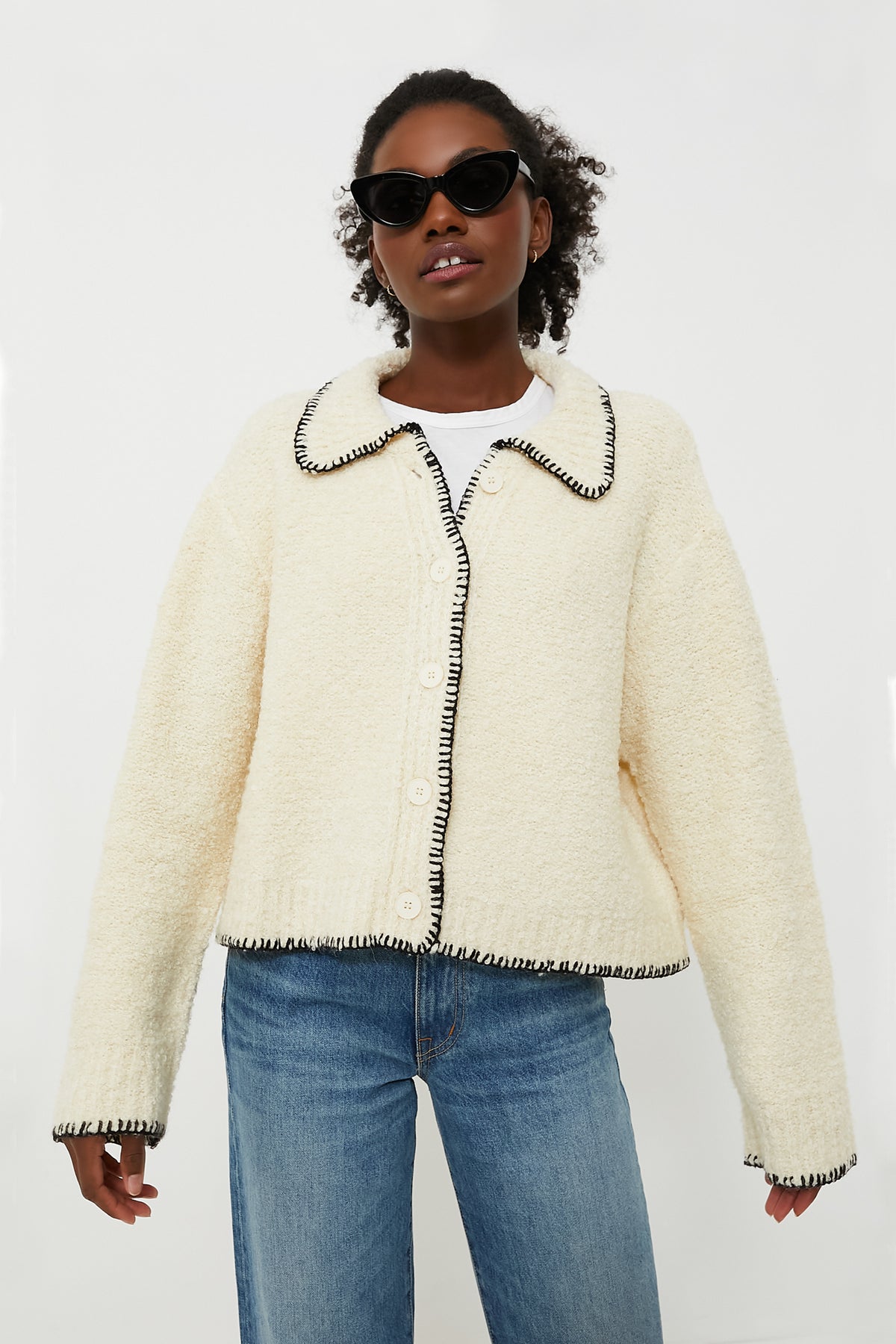 A person wearing black sunglasses, a cream Moon River cardigan with contrast stitching and black trim, and blue jeans stands confidently.
