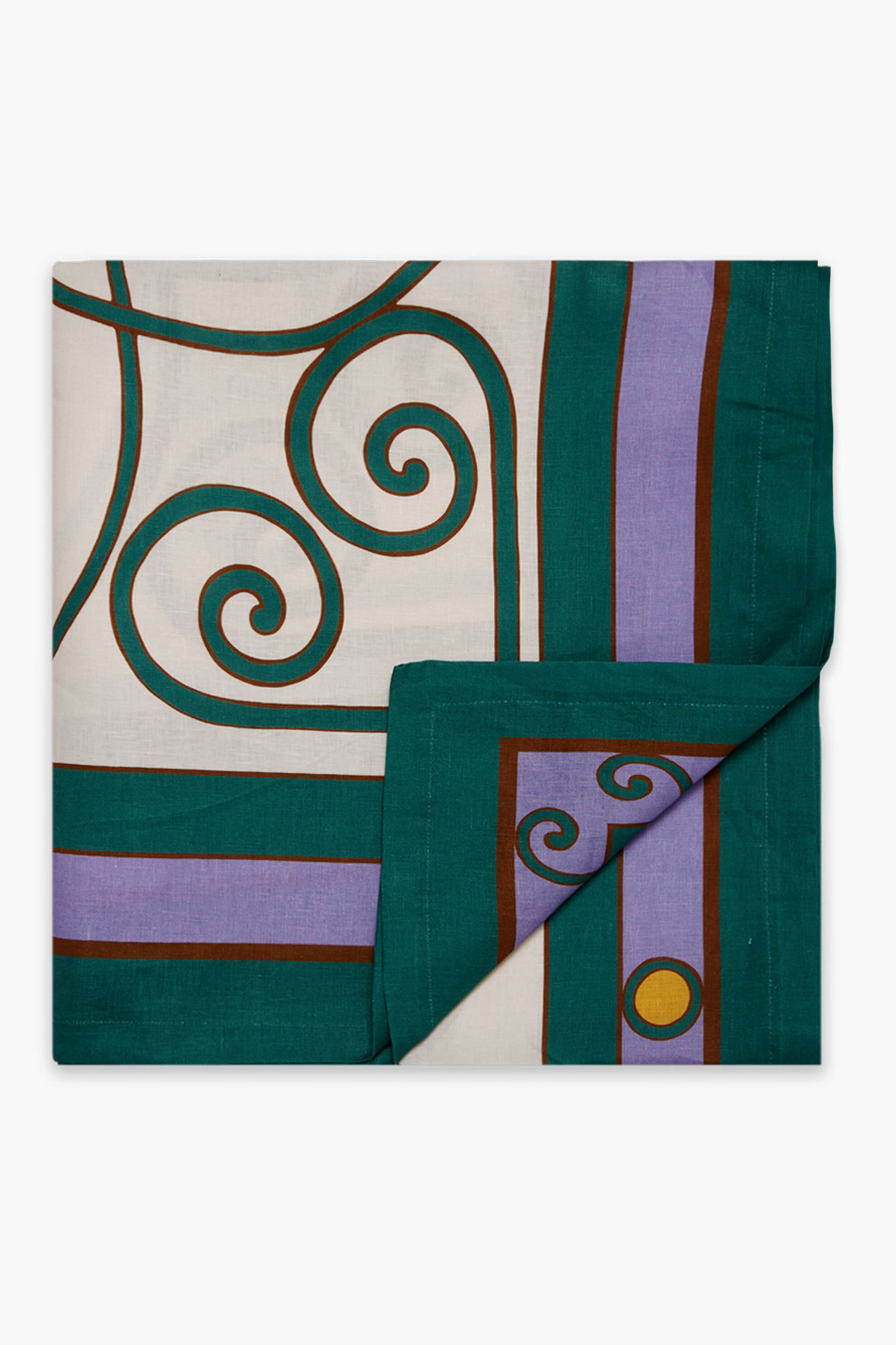 A folded La DoubleJ linen tablecloth with geometric and swirl patterns in teal, purple, brown, yellow, and white. A corner is turned up to display the underside of this large Italian-made tablecloth.