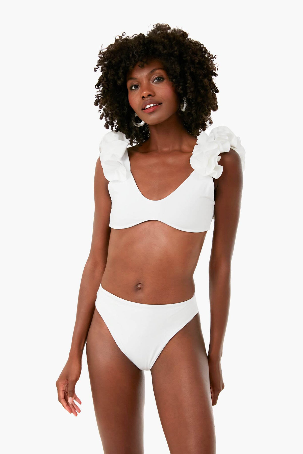 A woman wears a Maygel Coronel white swimsuit featuring flower-shaped ruffles on the shoulder straps and a high-rise leg.