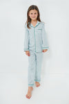 A young girl stands barefoot in Petite Plume’s kids cotton pajama set, featuring light blue and white stripes with green trim—an emerald ticking design made for cozy, restful nights.