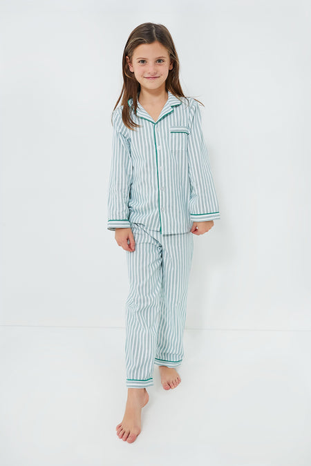 A young girl stands barefoot in Petite Plume’s kids cotton pajama set, featuring light blue and white stripes with green trim—an emerald ticking design made for cozy, restful nights.