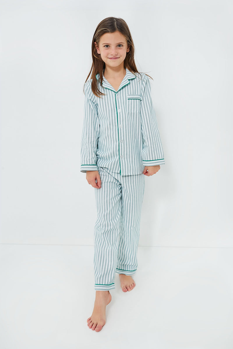 A young girl stands barefoot in Petite Plume’s kids cotton pajama set, featuring light blue and white stripes with green trim—an emerald ticking design made for cozy, restful nights.