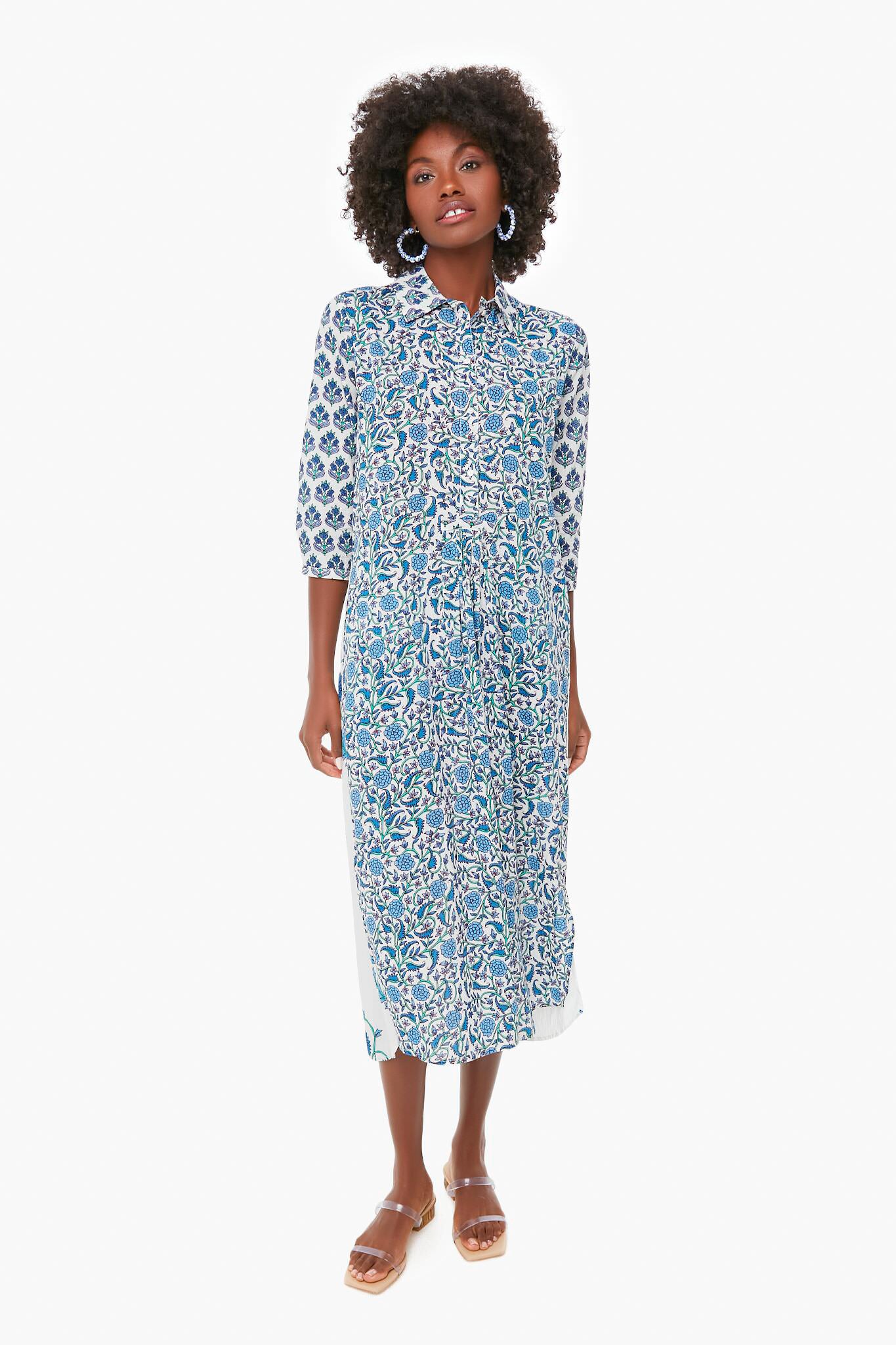 A woman wearing a Ro's Garden blue and white vibrant print midi dress with three-quarter sleeves, hoop earrings, and brown sandals—ideal luxury travel-wear for stylish getaways.