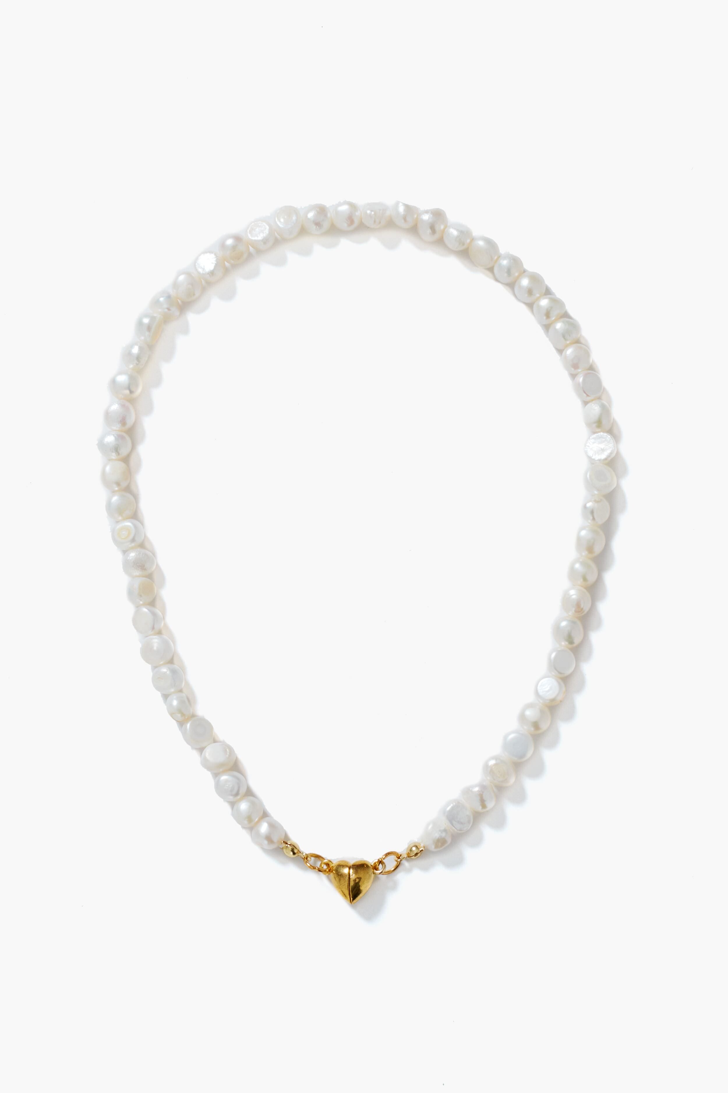 A Petit Moments white pearl necklace with gold-tone hardware and a heart-shaped magnetic closure, elegantly arranged in a circle.