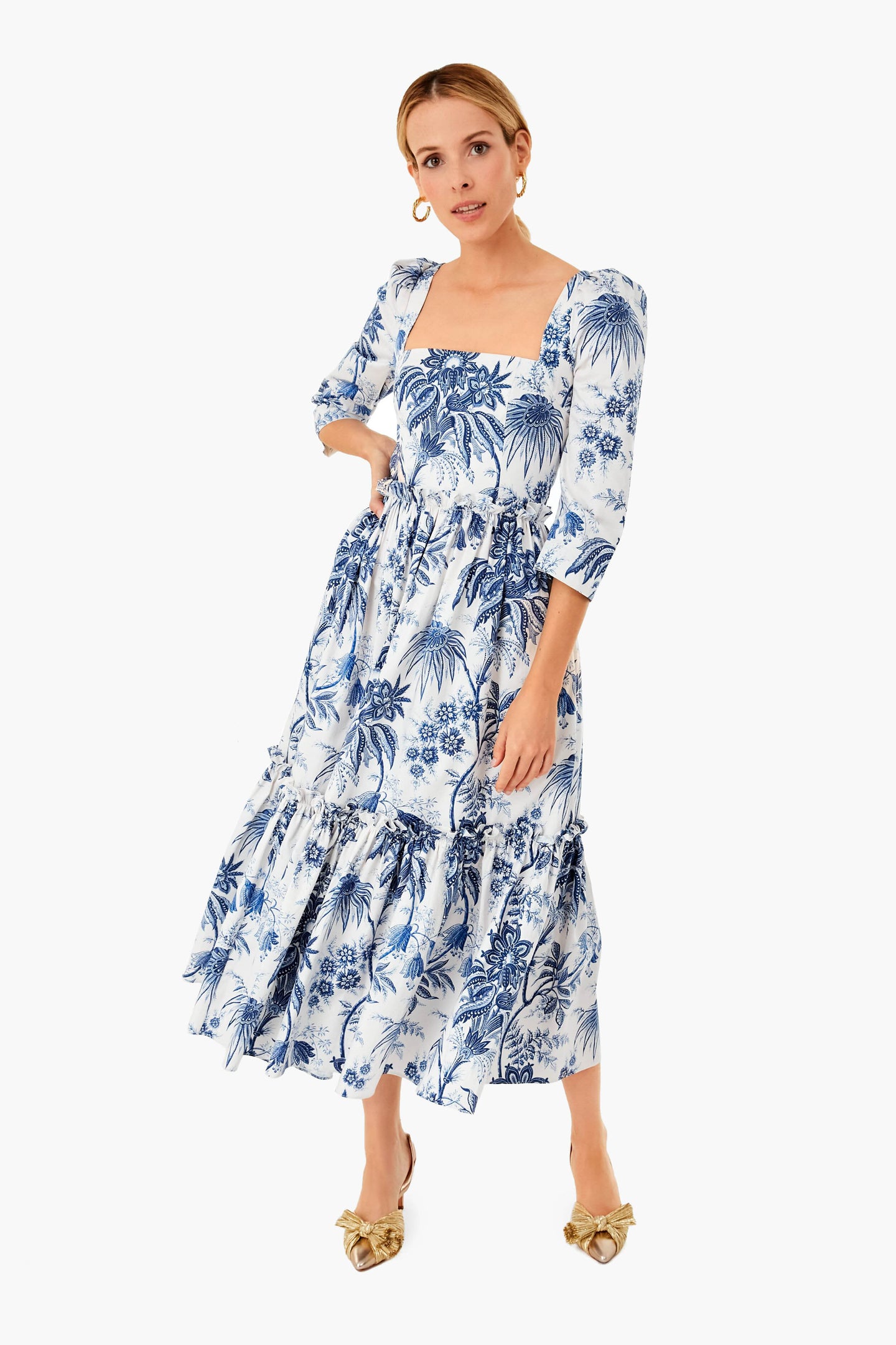 A woman wears a white and blue Jacobean toile cotton sateen midi dress with puffed sleeves by Cara Cara, paired with gold slip-on shoes.