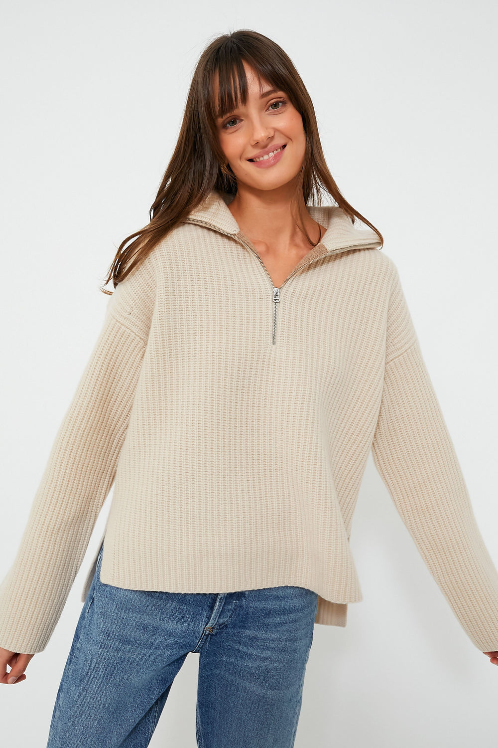 A woman smiles with arms slightly outstretched, wearing a beige ribbed zip mock neck sweater by Alex Mill and blue jeans.