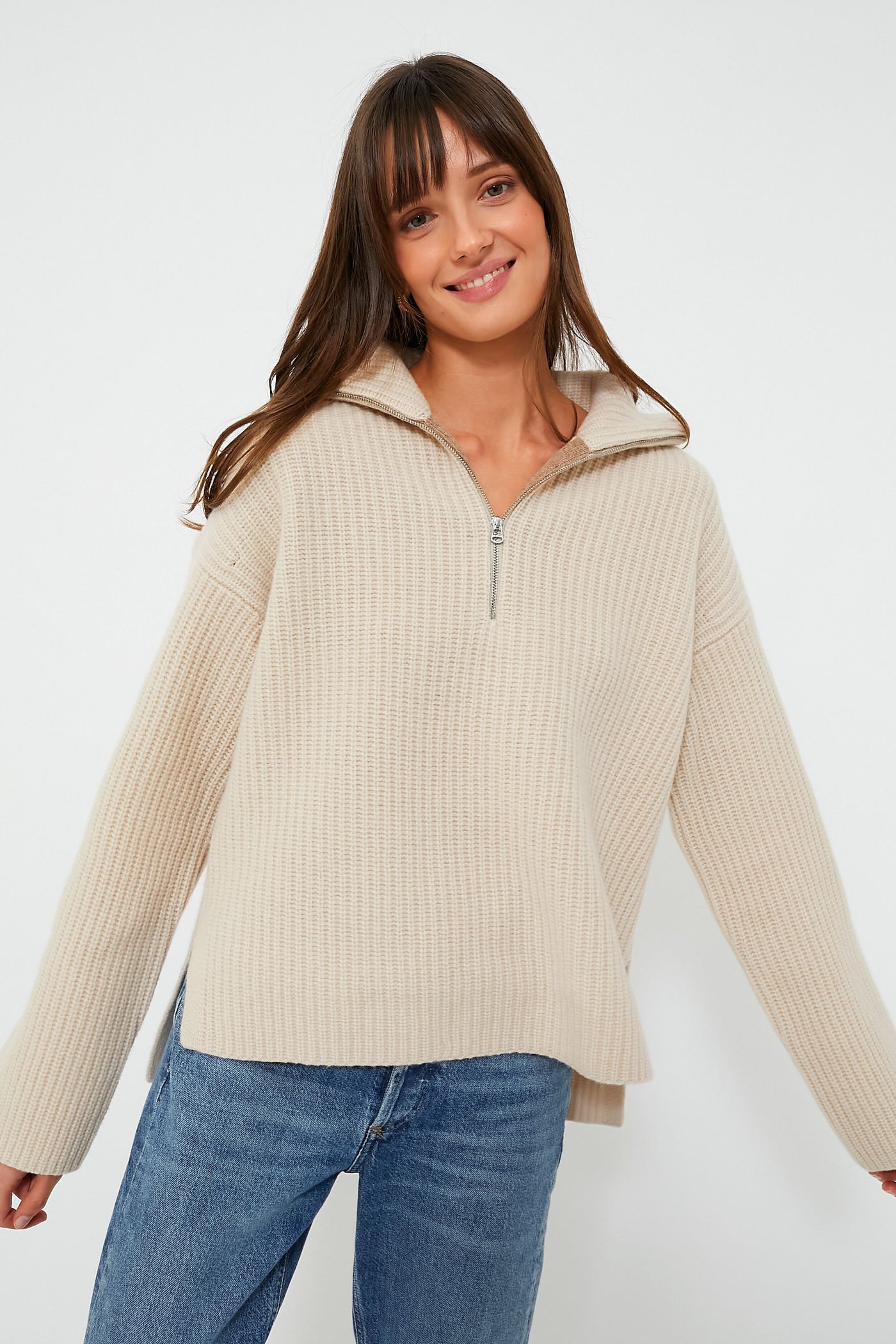 A woman smiles with arms slightly outstretched, wearing a beige ribbed zip mock neck sweater by Alex Mill and blue jeans.