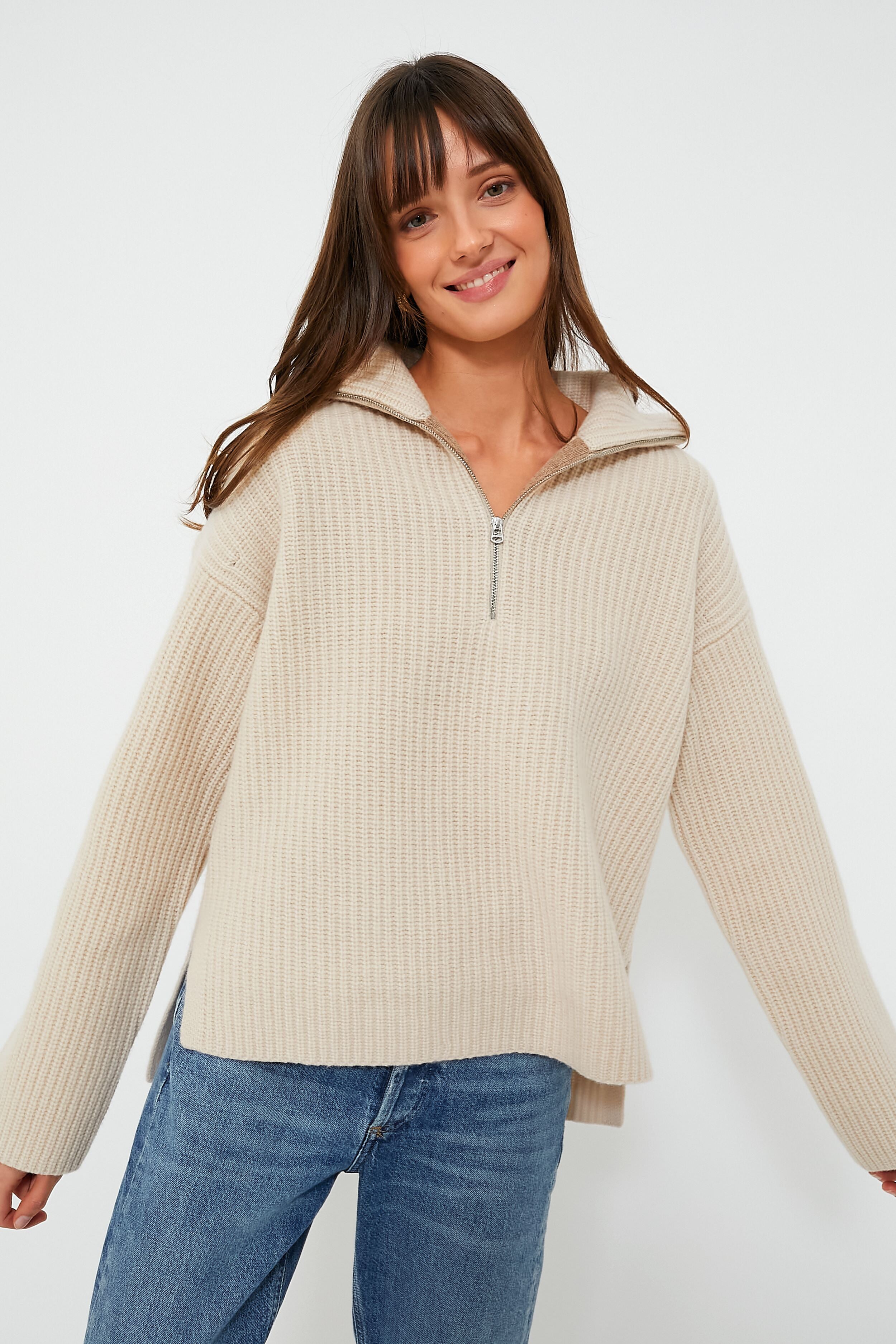A woman smiles with arms slightly outstretched, wearing a beige ribbed zip mock neck sweater by Alex Mill and blue jeans.