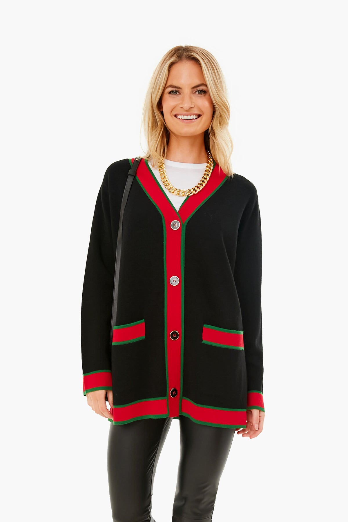A woman stands and smiles in a Tuckernuck oversized black cardigan with red and green trim, gold crest buttons, paired with a gold chain necklace, white shirt, and black pants. This wool piece adds chic style to any outfit.