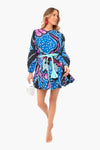 A woman wears a colorful, patterned blue RHODE Ella dress with a belted waist, holding a white clutch and accessorizing with nude sandals and bright pink earrings.