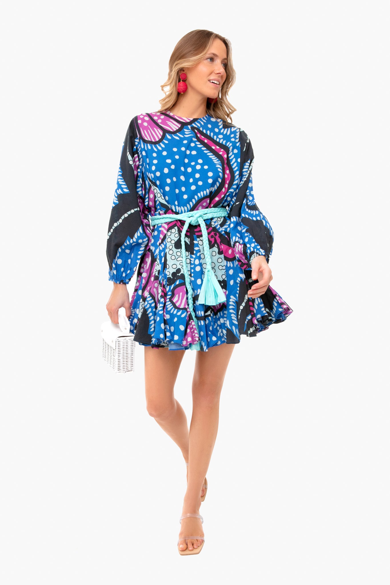 A woman wears a colorful, patterned blue RHODE Ella dress with a belted waist, holding a white clutch and accessorizing with nude sandals and bright pink earrings.