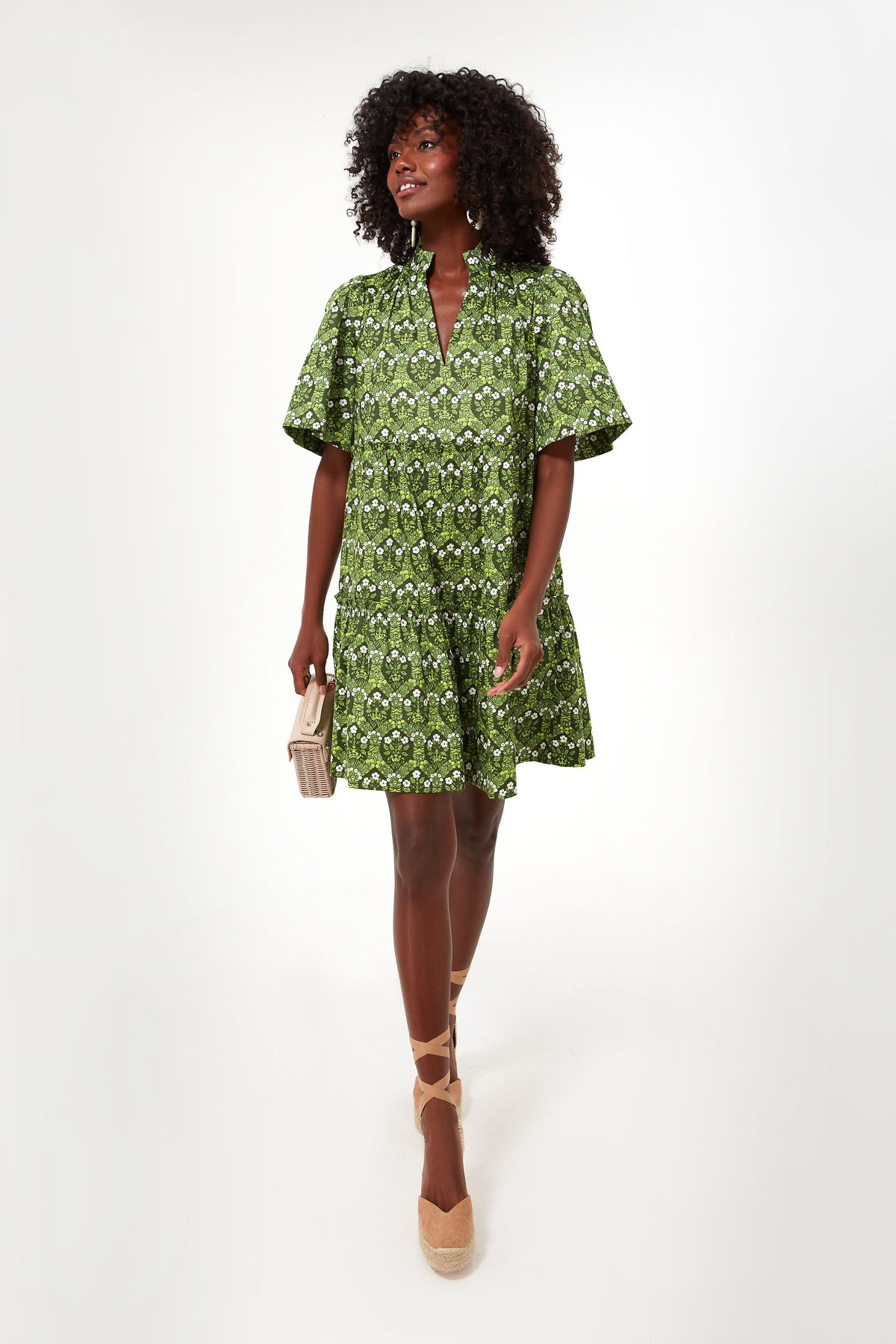 A woman stands wearing a Tuckernuck short-sleeved Crawford Dress with a vibrant spring print, tan wedge espadrilles, and holding a beige clutch.