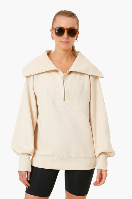 A person in Varley sunglasses, a beige half zip pullover with a wide collar, and black cycling shorts stands facing forward, blending performance fashion with casual style.