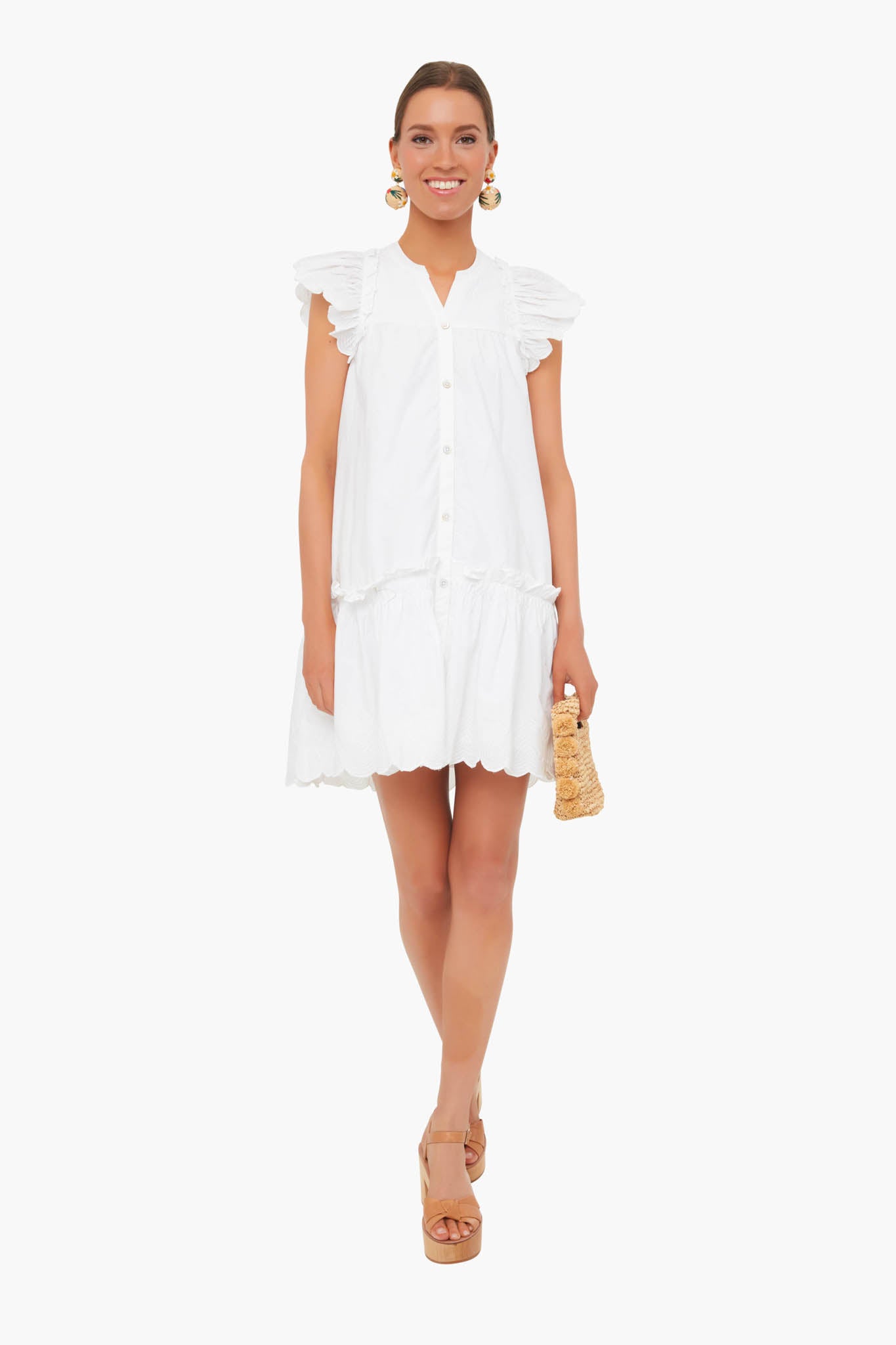 A woman wears a Sea New York white ruffled cotton dress with button-downs, tan platform sandals, large gold earrings, and holds a small woven clutch—perfect for resort style.