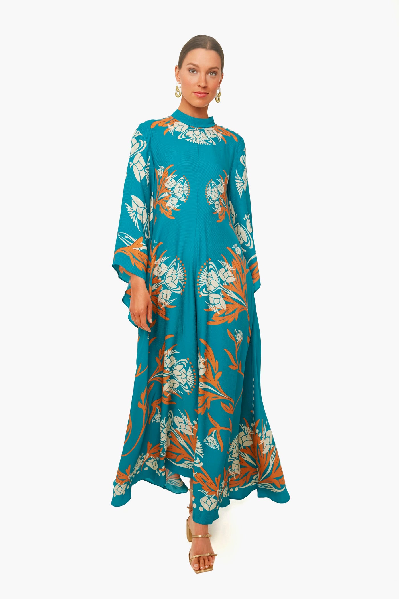 Woman in a La DoubleJ Florence Placee Magnifico Dress—teal maxi with orange and white floral print—paired with gold earrings and heels.