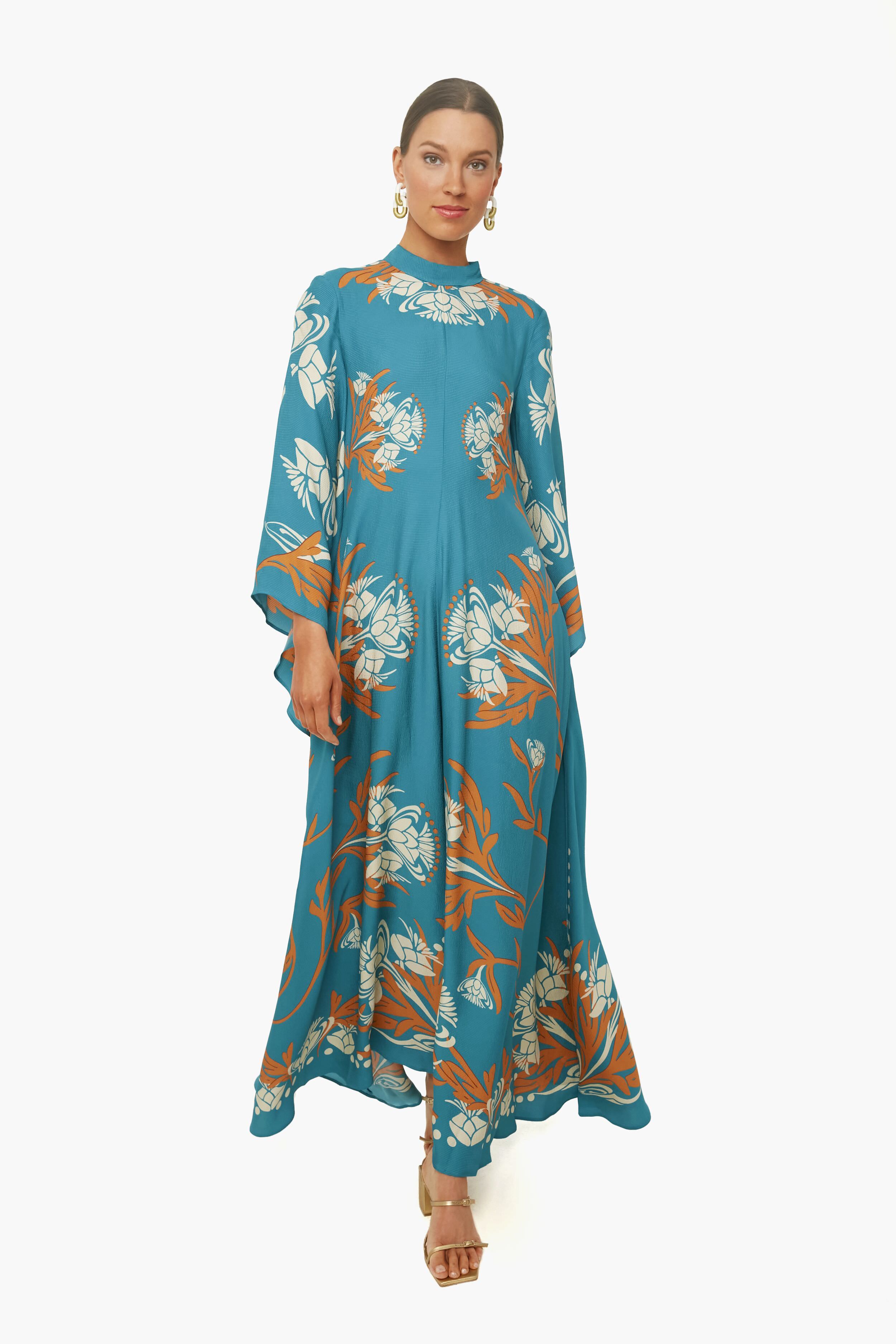 Woman in a La DoubleJ Florence Placee Magnifico Dress—teal maxi with orange and white floral print—paired with gold earrings and heels.