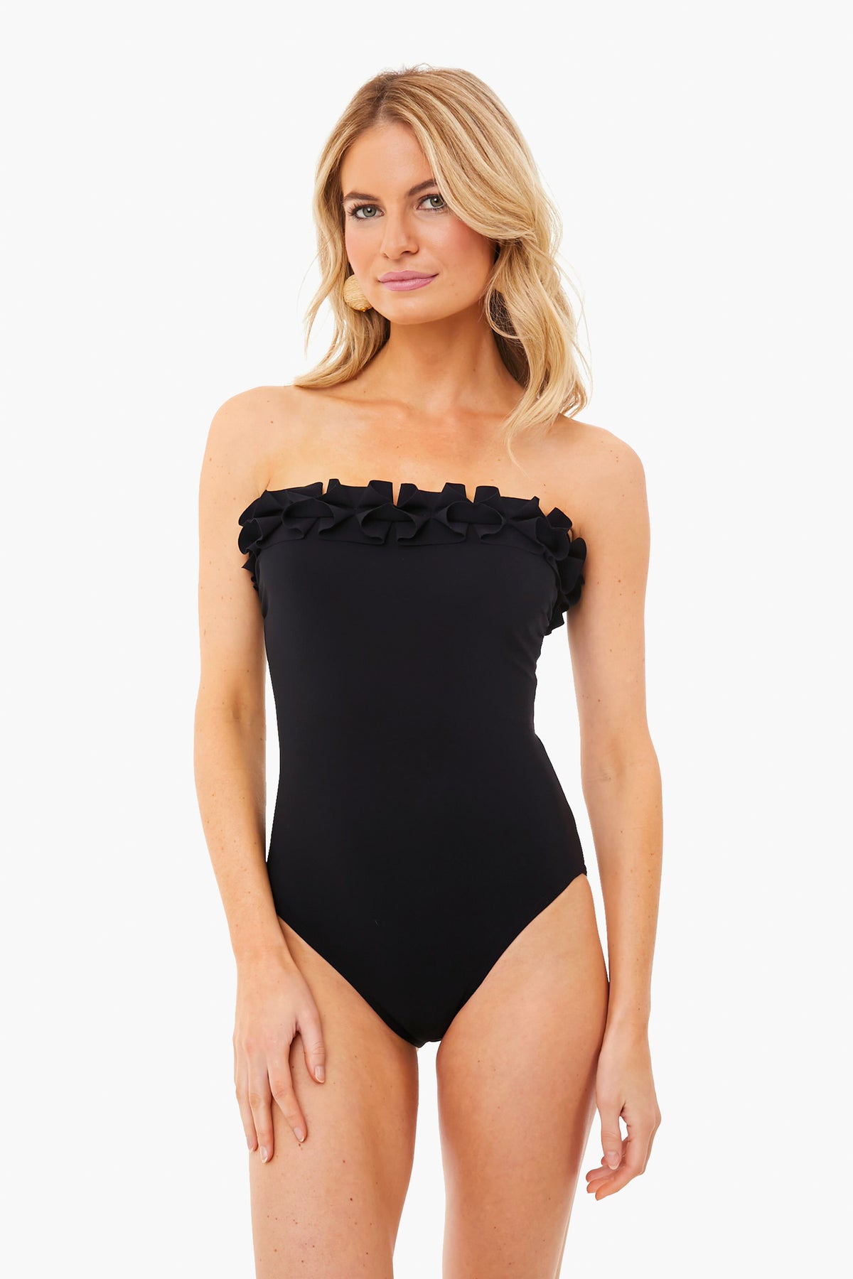 A woman stands confidently in a Karla Colletto black one-piece swimsuit featuring ruffle detail along the bandeau neckline.