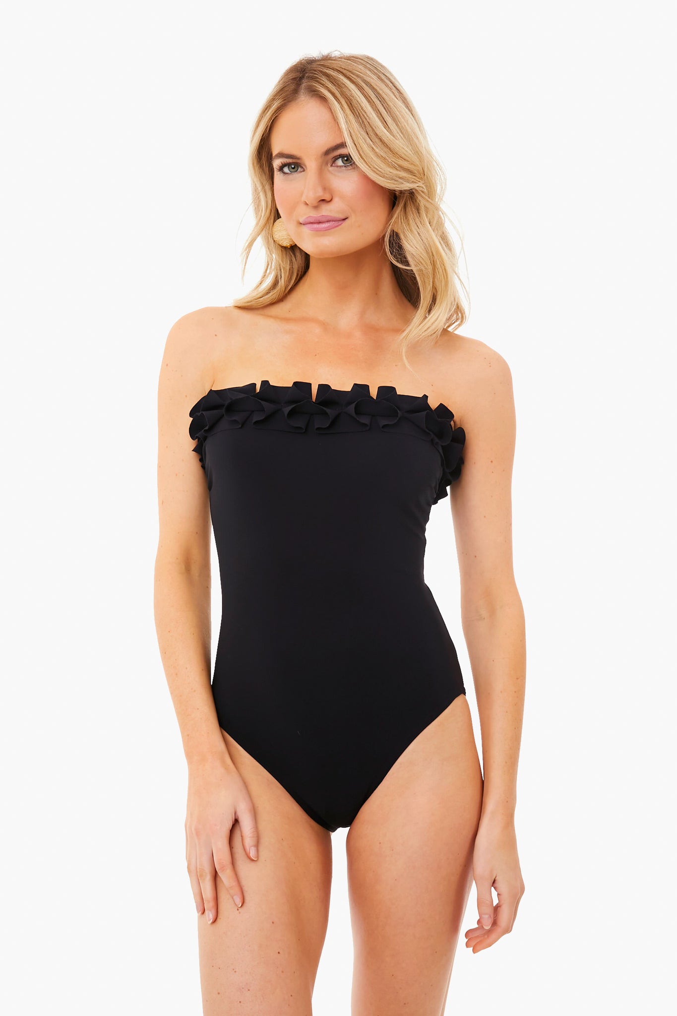 A woman stands confidently in a Karla Colletto black one-piece swimsuit featuring ruffle detail along the bandeau neckline.